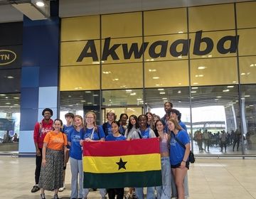 Participants of the 2024 High School Summer Abroad program take a photo in front of the Akwaaba sign at the Kotoka International Airport. A few of them standing in the front row hold up the Ghana national flag. 