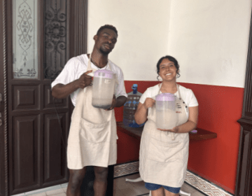 Two students wearing aprons stand side by side holding jars of water