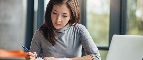 Woman writing in a notebook with her laptop open beside her