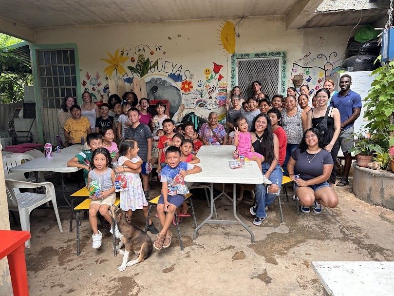 Group of children, teens and mothers stand and sit around tables, posing for a group photo. 