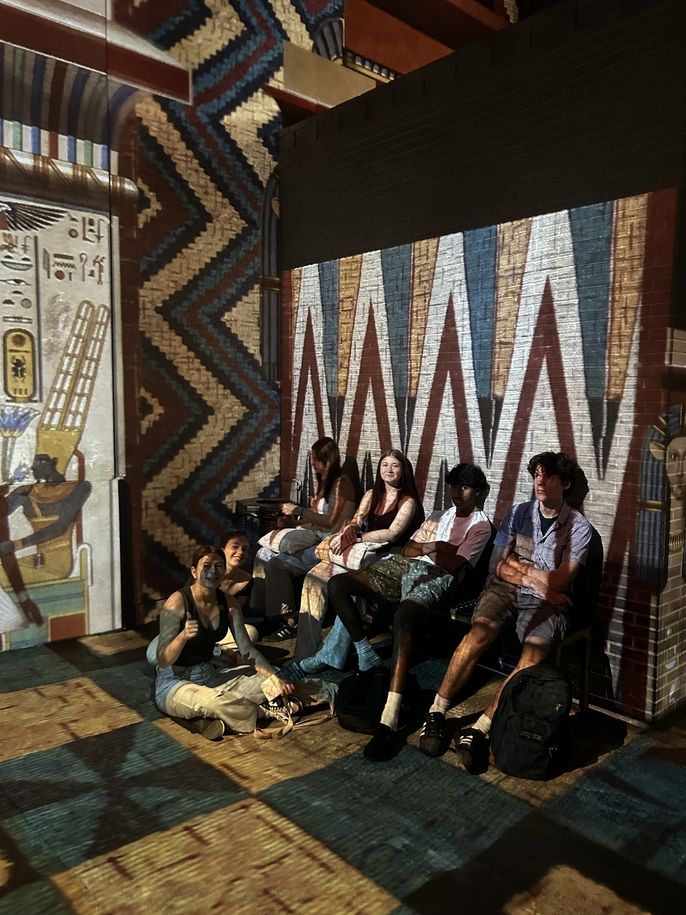 A group of HSSA Language and Culture students sit against a wall watching the light show. A zigzag mosaic pattern is projected on the wall behind them.