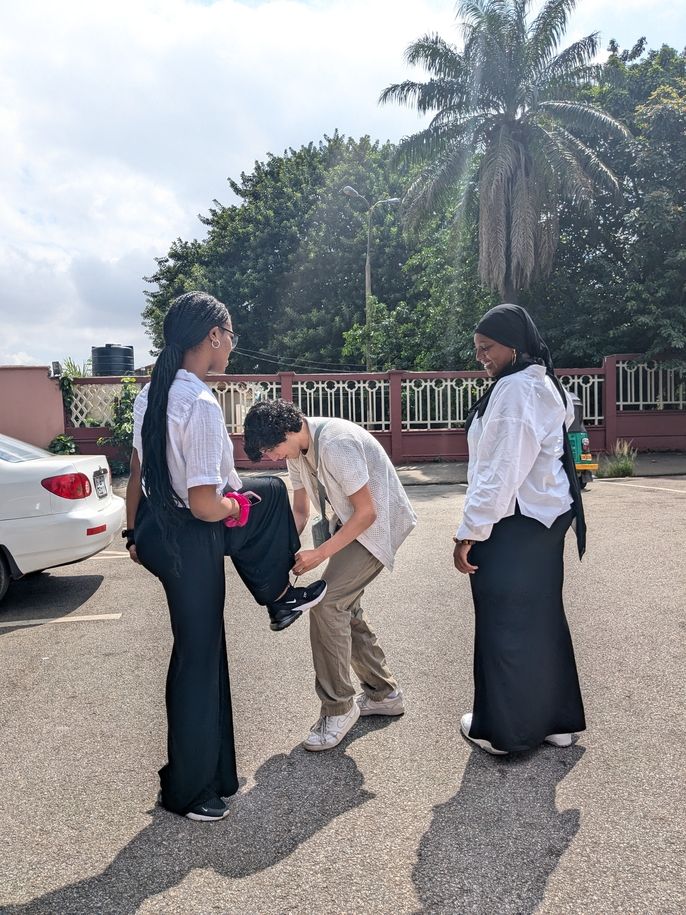 Max helps Shaiyanna lace her shoes. Shaiyanna has her left foot on Max's knee. Monajah looks on in the background.