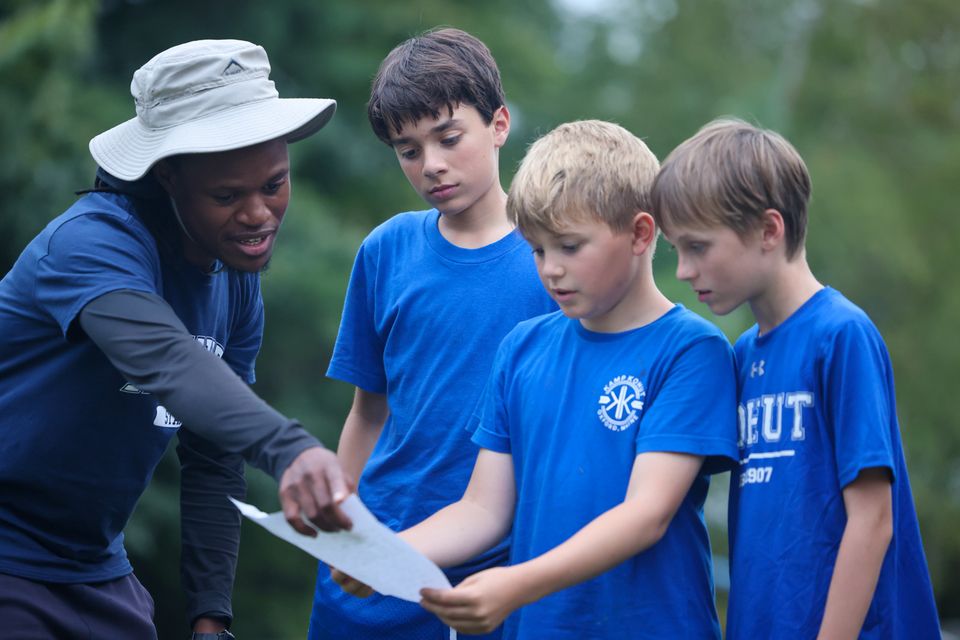 A close-up of a camp counselor wearing a wide-brimmed hat, pointing at a piece of paper or map, while three young boys in matching blue camp shirts attentively listen and look on.